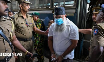 A man with a beard, skullcap and face mask being led out of a bus by four police officers