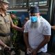 A man with a beard, skullcap and face mask being led out of a bus by four police officers