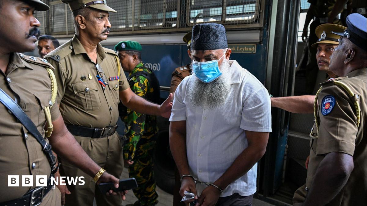 A man with a beard, skullcap and face mask being led out of a bus by four police officers