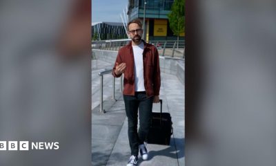 Consumer expert Harry Kind is walking, pulling a suitcase with his left hand. He's wearing a red jacket over a white t-shirt.