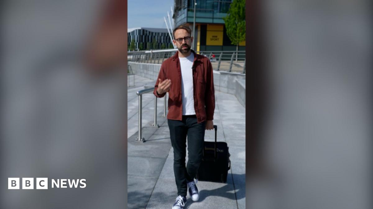 Consumer expert Harry Kind is walking, pulling a suitcase with his left hand. He's wearing a red jacket over a white t-shirt.