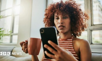 A woman looks at her phone with a mug in hand inside her home