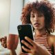 A woman looks at her phone with a mug in hand inside her home