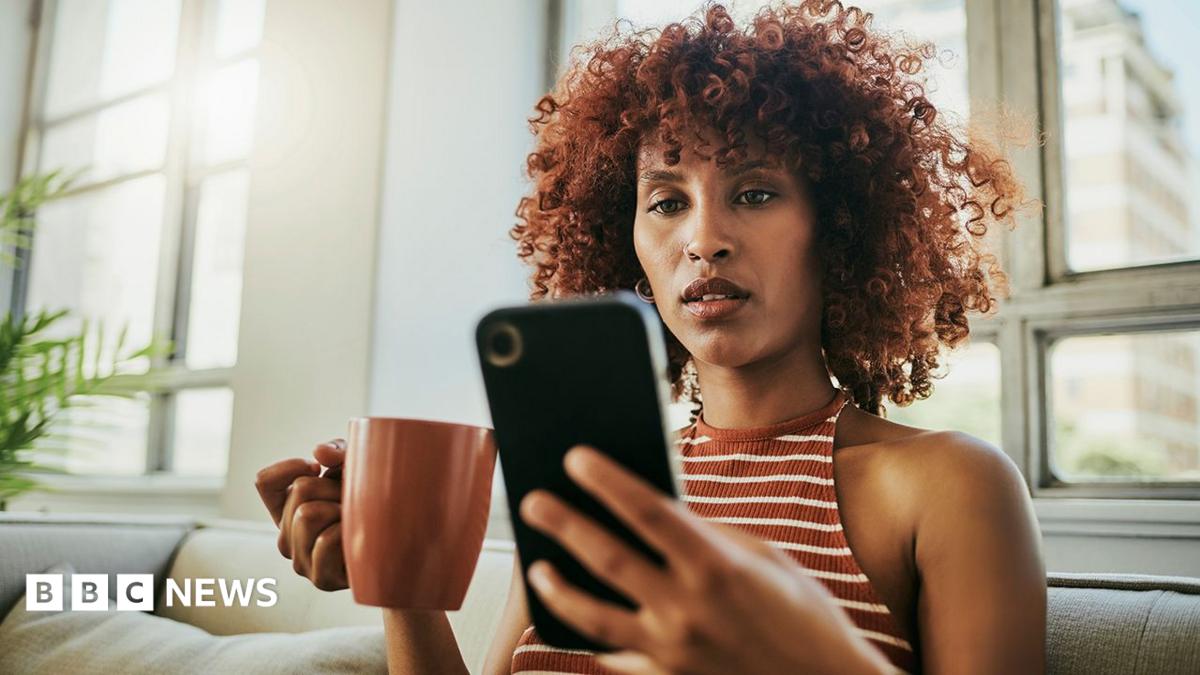 A woman looks at her phone with a mug in hand inside her home