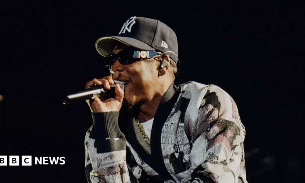 Jay-Z raps into a microphone while performing on stage. He wears a black and white cardigan and a New York Yankees hat.