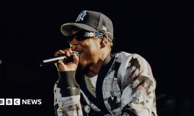 Jay-Z raps into a microphone while performing on stage. He wears a black and white cardigan and a New York Yankees hat.