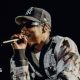 Jay-Z raps into a microphone while performing on stage. He wears a black and white cardigan and a New York Yankees hat.