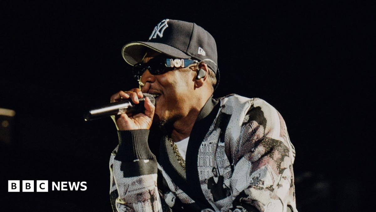 Jay-Z raps into a microphone while performing on stage. He wears a black and white cardigan and a New York Yankees hat.