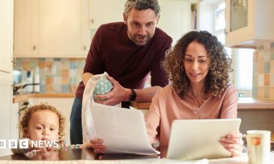 A family of three looking at bills and finances at a kitchen table. From left to right, a child is standing on the floor looking out across the table, a man is looking over a woman's shoulder at A4 sheets of paper she is holding while standing drying a mug, and a woman sits at the table holding the papers and reading. The kitchen behind them in soft focus is mostly pastel-coloured.