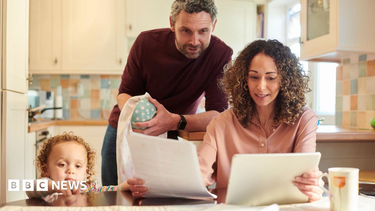 A family of three looking at bills and finances at a kitchen table. From left to right, a child is standing on the floor looking out across the table, a man is looking over a woman's shoulder at A4 sheets of paper she is holding while standing drying a mug, and a woman sits at the table holding the papers and reading. The kitchen behind them in soft focus is mostly pastel-coloured.