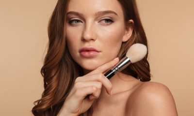 Woman with auburn hair and natural makeup holding a powder brush near her jaw against a warm beige background
