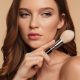 Woman with auburn hair and natural makeup holding a powder brush near her jaw against a warm beige background
