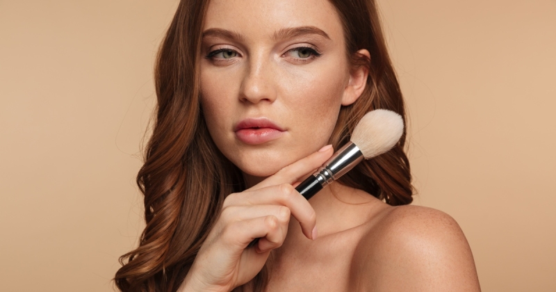 Woman with auburn hair and natural makeup holding a powder brush near her jaw against a warm beige background