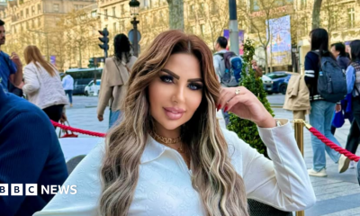 Sarah Khalifa wearing a white long-shirted with her folded left arm, the hand touching her long hair, sits by the side of a road inside a red-roped cordoned area, beyong which people walk on a road the other side of which has rows of sunbathed buildings
