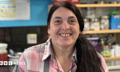 Patsy Lockerby, who has long dark hair in the ponytail and is wearing a pink, white and grey checked shirt. She'd standing behind the counter in her tearoom.