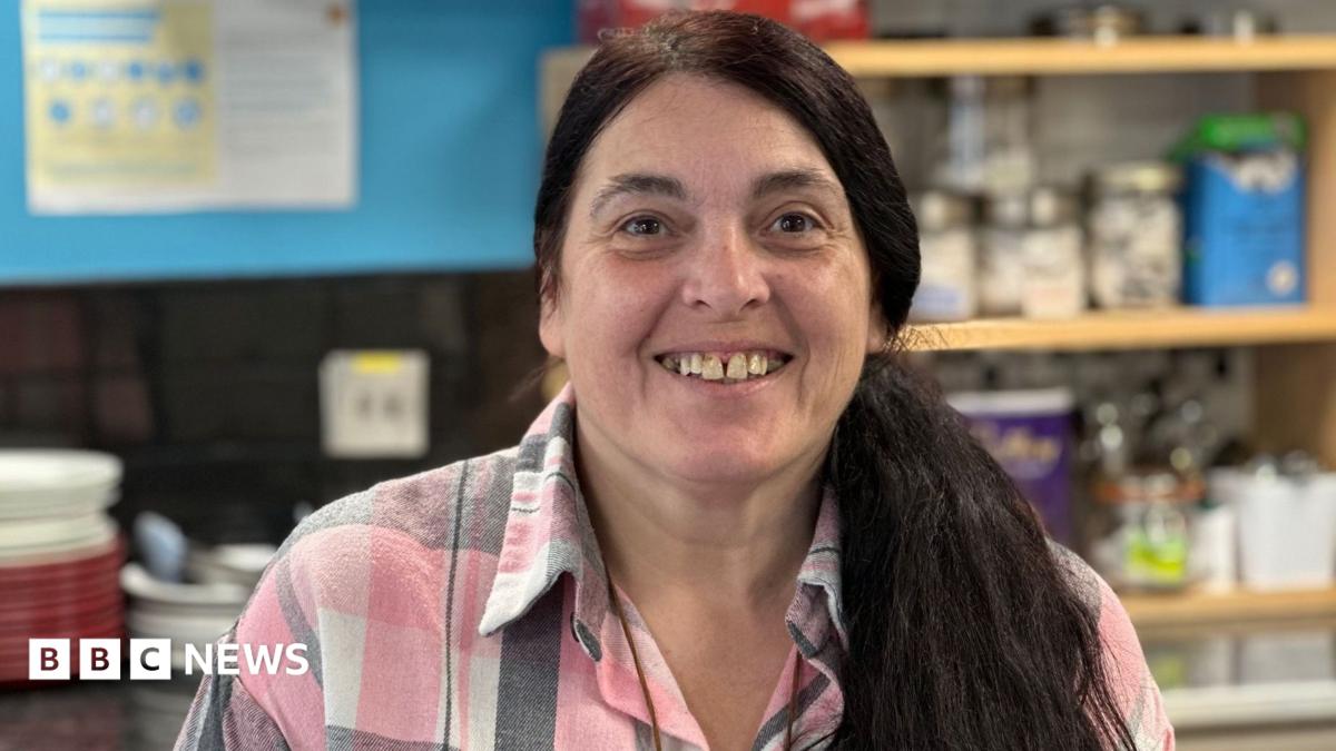 Patsy Lockerby, who has long dark hair in the ponytail and is wearing a pink, white and grey checked shirt. She'd standing behind the counter in her tearoom.
