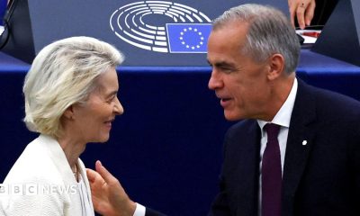 A smiling Mark Carney, wearing a suit, walks alongside European Parliament President Roberta Metsola and Ursula von der Leyen at the European Parliament
