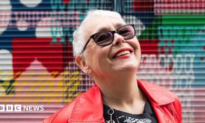 A tightly cropped photo of a woman in her 70s with short grey hair looking up at the sky, smiling. She has sunglasses on, and bright red lipstick that matches her leather jacket. Graffiti is in the background, slightly out of focus.