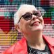 A tightly cropped photo of a woman in her 70s with short grey hair looking up at the sky, smiling. She has sunglasses on, and bright red lipstick that matches her leather jacket. Graffiti is in the background, slightly out of focus.