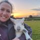 A person is taking a selfie while holding a young lamb in a grassy field at sunset. The lamb is white with dark markings on its nose and legs, and in the background there are several sheep scattered across green pasture, hedgerows, trees and a colourful orange and blue evening sky stretching across the horizon.