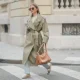 Diane Batoukina wears her long straight brown hair down with a center part, oversized square Dior sunglasses, a khaki belted trench coat by Sandro layered over a beige T-shirt by Zara, light-wash straight-leg jeans by Anine Bing, beige suede Miu Miu sneakers with mismatched laces and tassel details, and carries a large tan leather shoulder bag by Prada, walking across a pedestrian crossing, during a street style fashion photo session on March 25, 2025 in Paris, France.