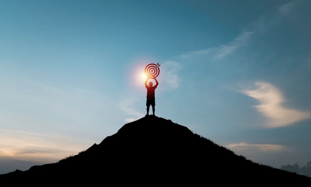 Silhouette of man holding dartboard with arrow on the top of mountain and blue sky for business achievement objective target and goal concept.