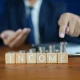 Businessman Presenting Income Growth with Stacked Coins on Wooden Table and Focused Gesture in Office Environment