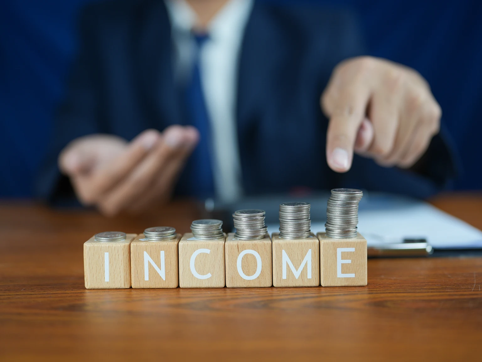 Businessman Presenting Income Growth with Stacked Coins on Wooden Table and Focused Gesture in Office Environment
