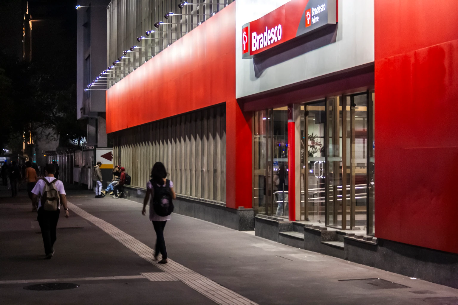 Night view of a Bradesco bank branch facade on Avenida Paulista. Pedestrians walk on the illuminated tactile sidewalk in the main financial district.