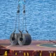 Cargo is being lifted from a ship above the ocean with cranes in a busy port during a sunny day