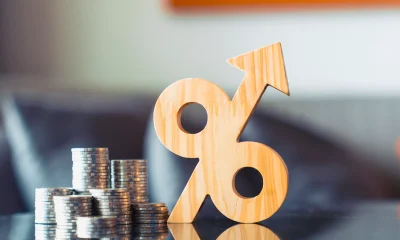Wooden percentage symbol with upward arrow next to growing coin stacks. Financial interest rate hike, rising inflation, profitable return, and tax growth concept.