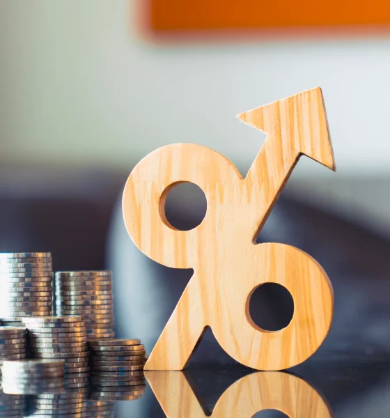 Wooden percentage symbol with upward arrow next to growing coin stacks. Financial interest rate hike, rising inflation, profitable return, and tax growth concept.