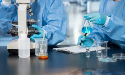 Scientists in protective clothing conduct laboratory research with a microscope, glassware, blue liquid sample and notes on a clean workbench.
