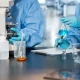 Scientists in protective clothing conduct laboratory research with a microscope, glassware, blue liquid sample and notes on a clean workbench.