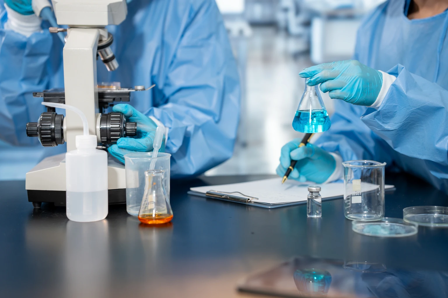 Scientists in protective clothing conduct laboratory research with a microscope, glassware, blue liquid sample and notes on a clean workbench.