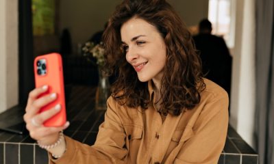 Smiling woman with curly hair in a camel shirt taking a selfie with an iPhone in a bright orange case