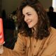 Smiling woman with curly hair in a camel shirt taking a selfie with an iPhone in a bright orange case