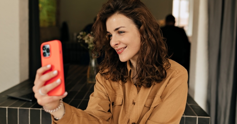 Smiling woman with curly hair in a camel shirt taking a selfie with an iPhone in a bright orange case