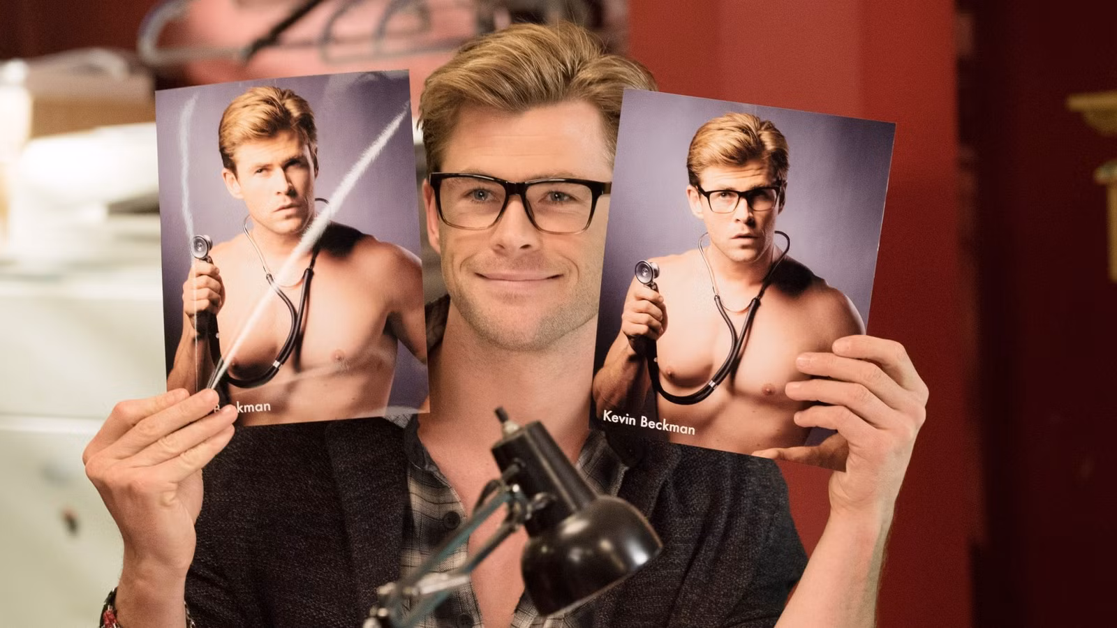 Chris Hemsworth sits at a desk holding up 2 almost identical photos of himself in Ghostbusters 2016