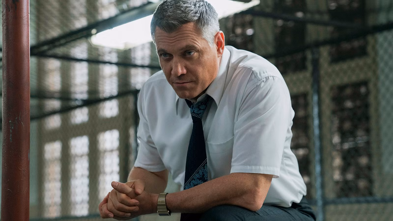 Bill leaning forward in front of a chain link barrier in 'Mindhunter'