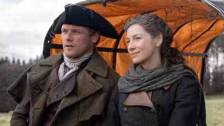 Sam Heughan as Jamie sitting on the back of a wagon next to Caitriona Balfe as Claire in Outlander Season 8
