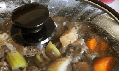 a crockpot slow cooker has the lid on with a beef stew cooking inside; in the background are red tomatoes and a blue and green kitchen towel