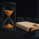Editorial illustration of an hourglass with draining sand beside a stack of sealed documents on a hearing room table, symbolizing a rulemaking deadline approaching