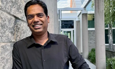 an indian man in a dress shirt smiling in front of the entrance to an office building