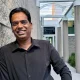 an indian man in a dress shirt smiling in front of the entrance to an office building