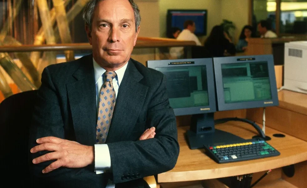 color photo of a white man in a business suit posing in front of a computer with office workers in t