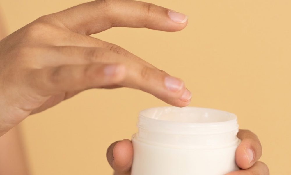woman dips her finger into a pot of moisturizer or cream; there is a very orangey background