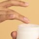 woman dips her finger into a pot of moisturizer or cream; there is a very orangey background